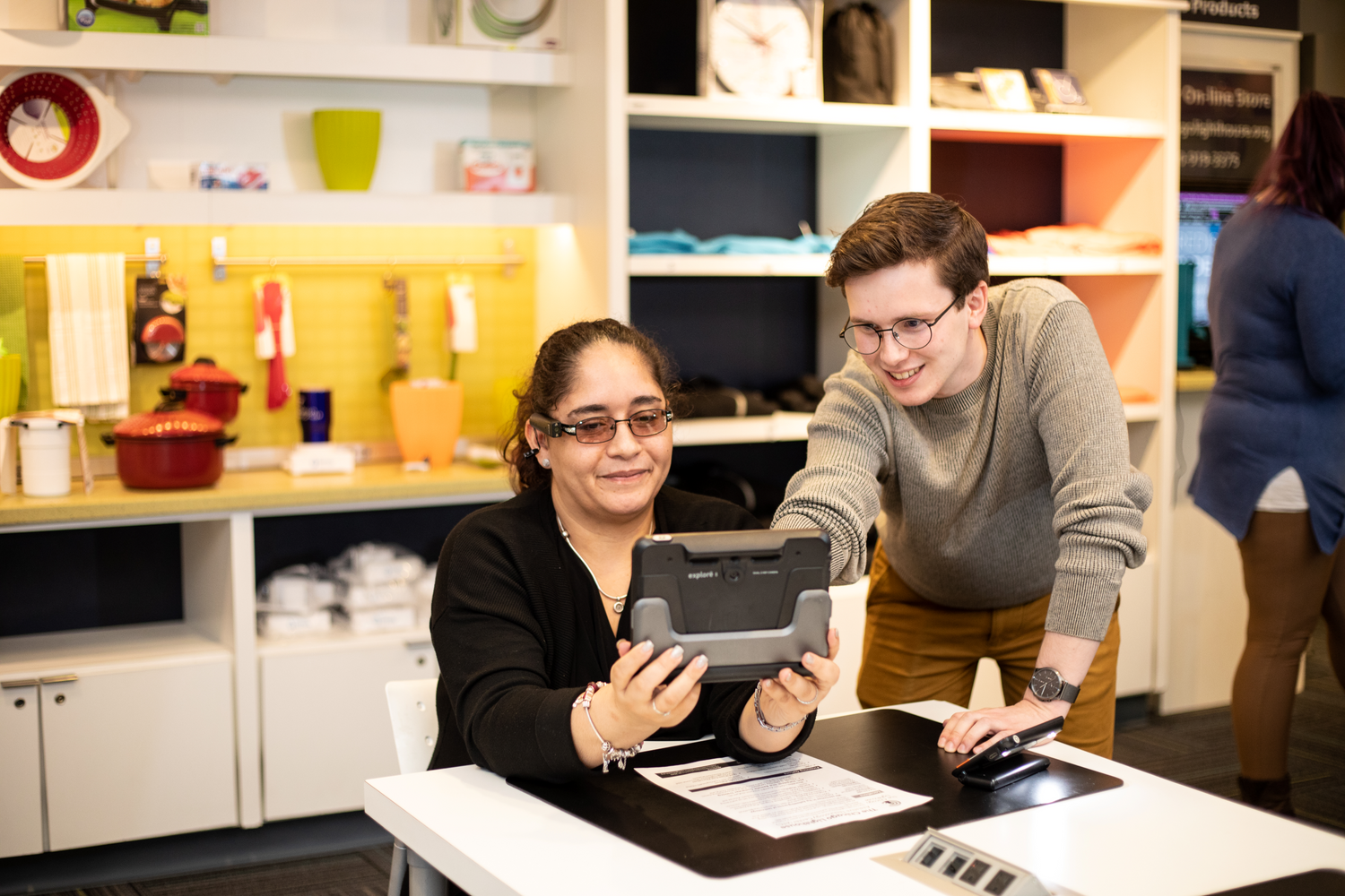 A woman sitting at a table holding a Humanware Explore 8 and wearing an OrCam MyEye device. A man is standing on her left side pointing towards the screen of the Humanware device.