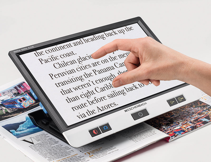 Hand using the touchscreen on the Eschenbach Visolux XL HD to magnify printed magazine text.