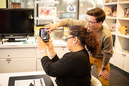 A woman sitting holding the Humanware Explore 8, magnifying into the distance while a man leans over her shoulder, touching the screen of the device.