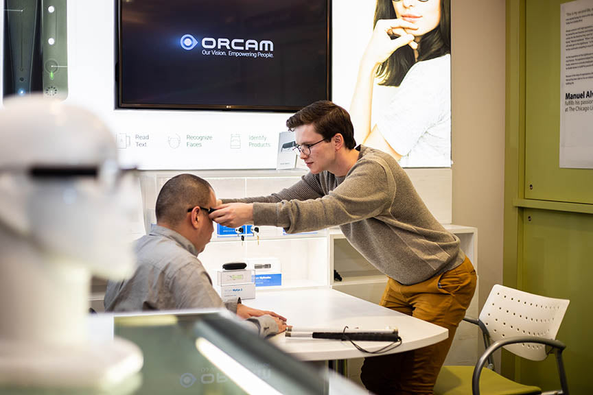 At the forefront & out of focus is a mannequin head wearing an OrCam device. In focus behind it are two men sitting at a table facing one another. The man seated on the left is having glasses put on him by the other man. The OrCam logo is behind them.