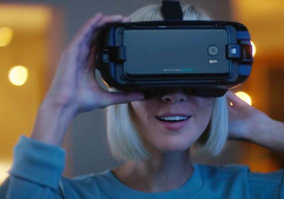 A young blonde woman smiling while using the IrisVision Live electronic glasses. The esight glasses are in a black color that resembles a Virtual Reality device.