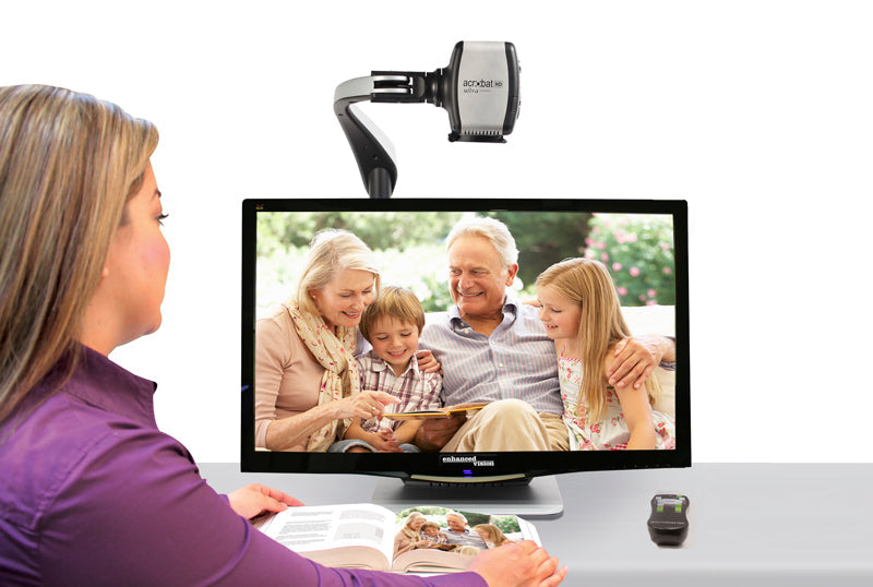 A woman sits in front of the Acrobat HD Ultra 27"  screen. The 3-1 camera attached to a metal arm above the screen is margnifing the image of a family of four reading a book.  A black remote with white and green button sits to the right of the open book