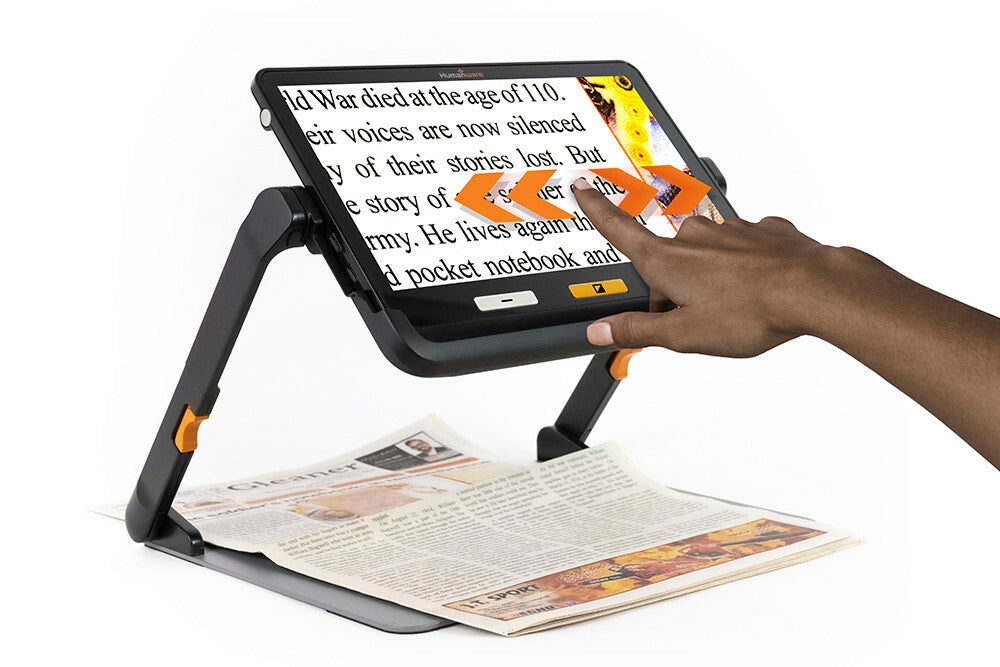 Humanware Explore 12 in it's stand showing a hand touching the touch screen while enlarging a newspaper underneath the device.