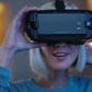 A young blonde woman smiling while using the IrisVision Live electronic glasses. The esight glasses are in a black color that resembles a Virtual Reality device. 