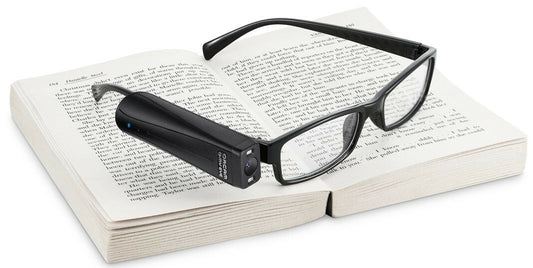 Orcam Myeye electronic glasses in black laying on top of an open book on a white background. The glasses are stylish bold frames. On the right side of the glasses is what looks like a clipped on black device that is the brains of the device that has an optical sensor.