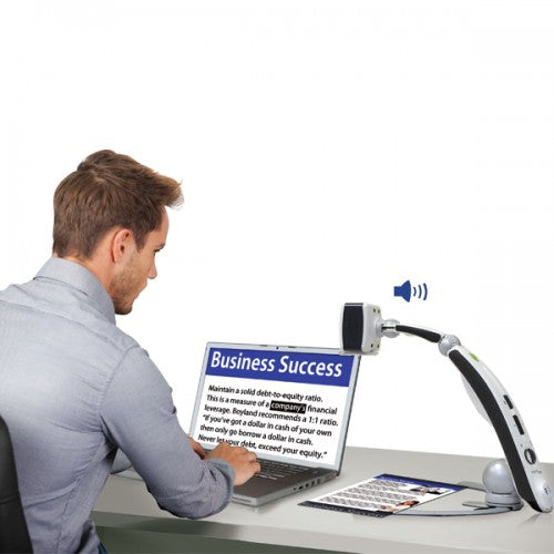 A gentleman seated at a desk is looking at his laptop screen with the Transformer HD electronic magnifier on the right side of the desk. The transformer HS camera is pointed down on a document that is showing the image on his computer screen that reads "business success" and other words below. A microphone sound above the device indicated that the document is being read out loud   